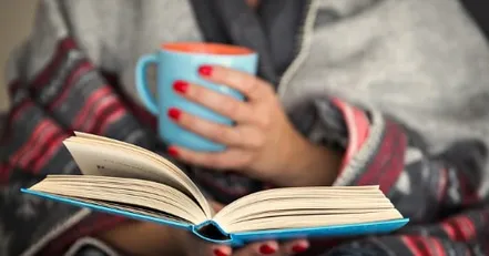 A mother reading a book and holding a mug of coffee"