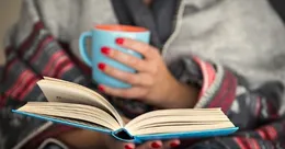 A mother reading a book and holding a mug of coffee"