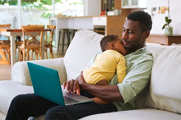 Father holding sleeping baby reading messages on his laptop